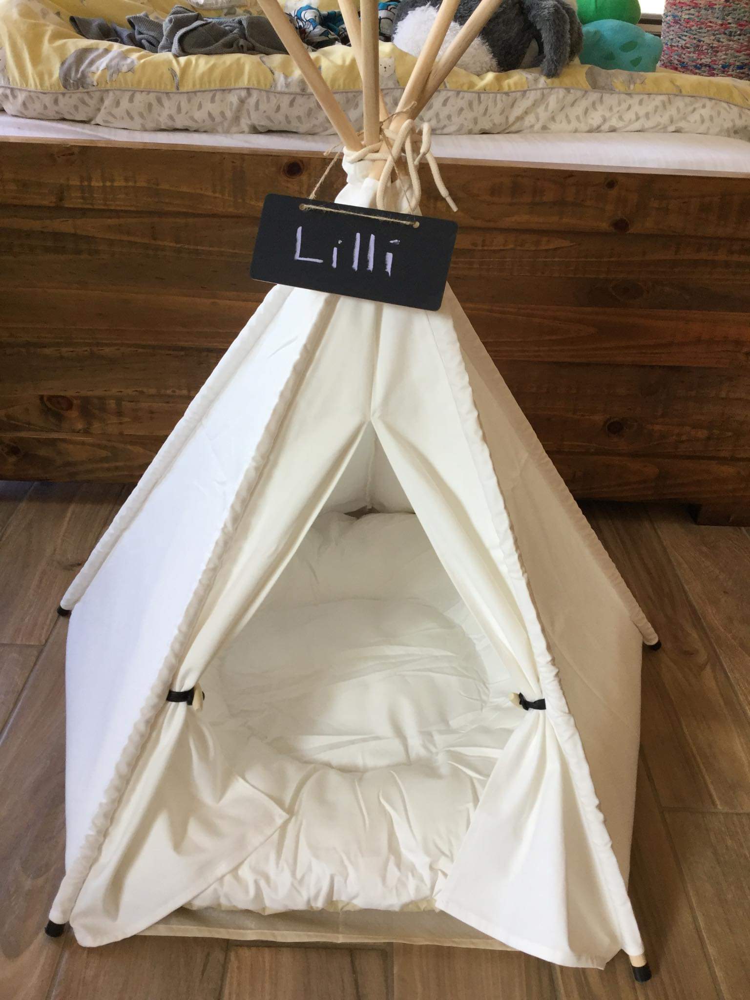 A white teepee tent designed specifically for cats that's placed near a bed, complete with a sign depicting the cat's name.